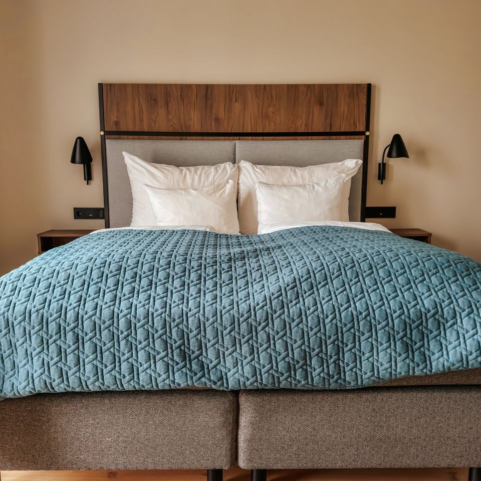 A hotel bed with turquoise quilted bed linen, white cushions and a headboard made of wood and grey fabric, flanked by two black wall lamps.