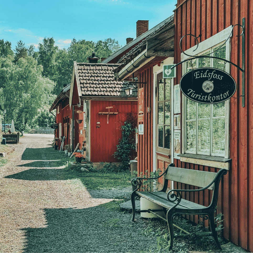 A row of red wooden houses along a gravel footpath, with a wrought-iron bench in front.
