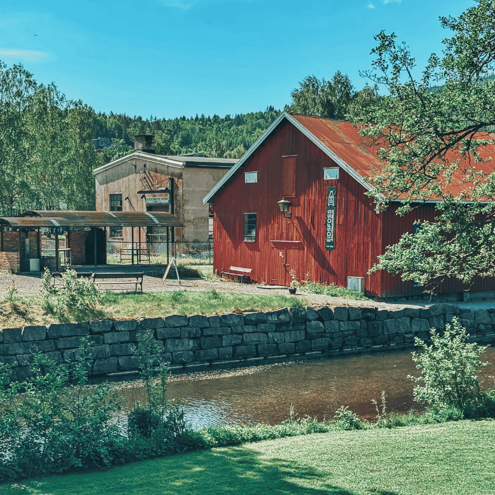 A red wooden house by a canal, with a stone wall, a lawn and a bench in the foreground.