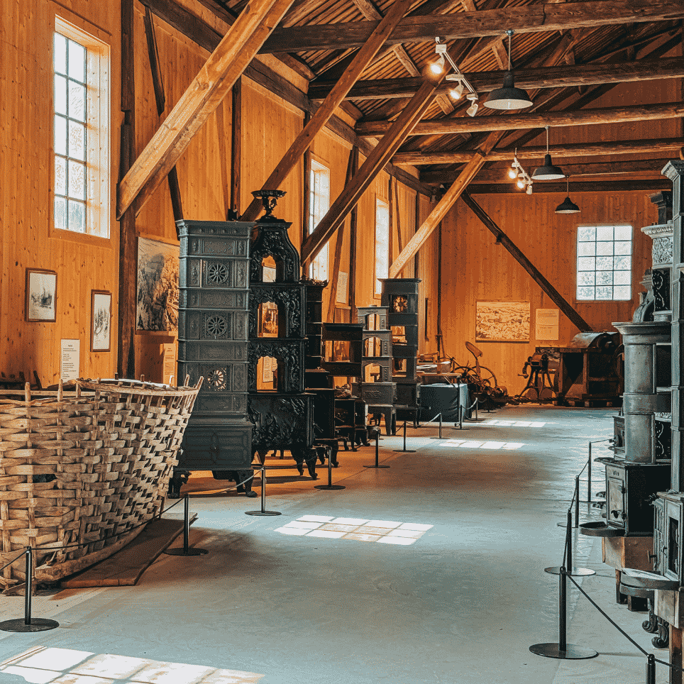 A museum exhibition featuring old iron stoves and a woven basket, beneath a beamed ceiling.
