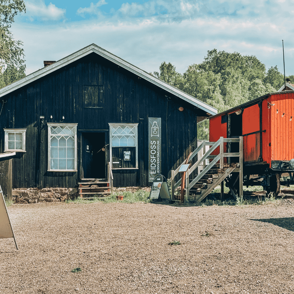 A dark wooden house with a museum sign, next to a red railway carriage on a gravel surface.