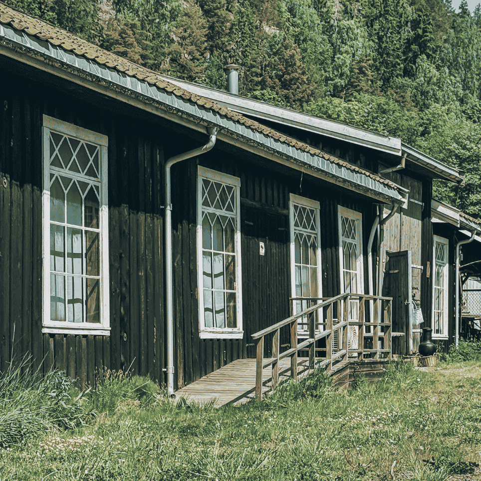 An old wooden house with white window frames and a wooden ramp leading up to the front door, surrounded by woodland.