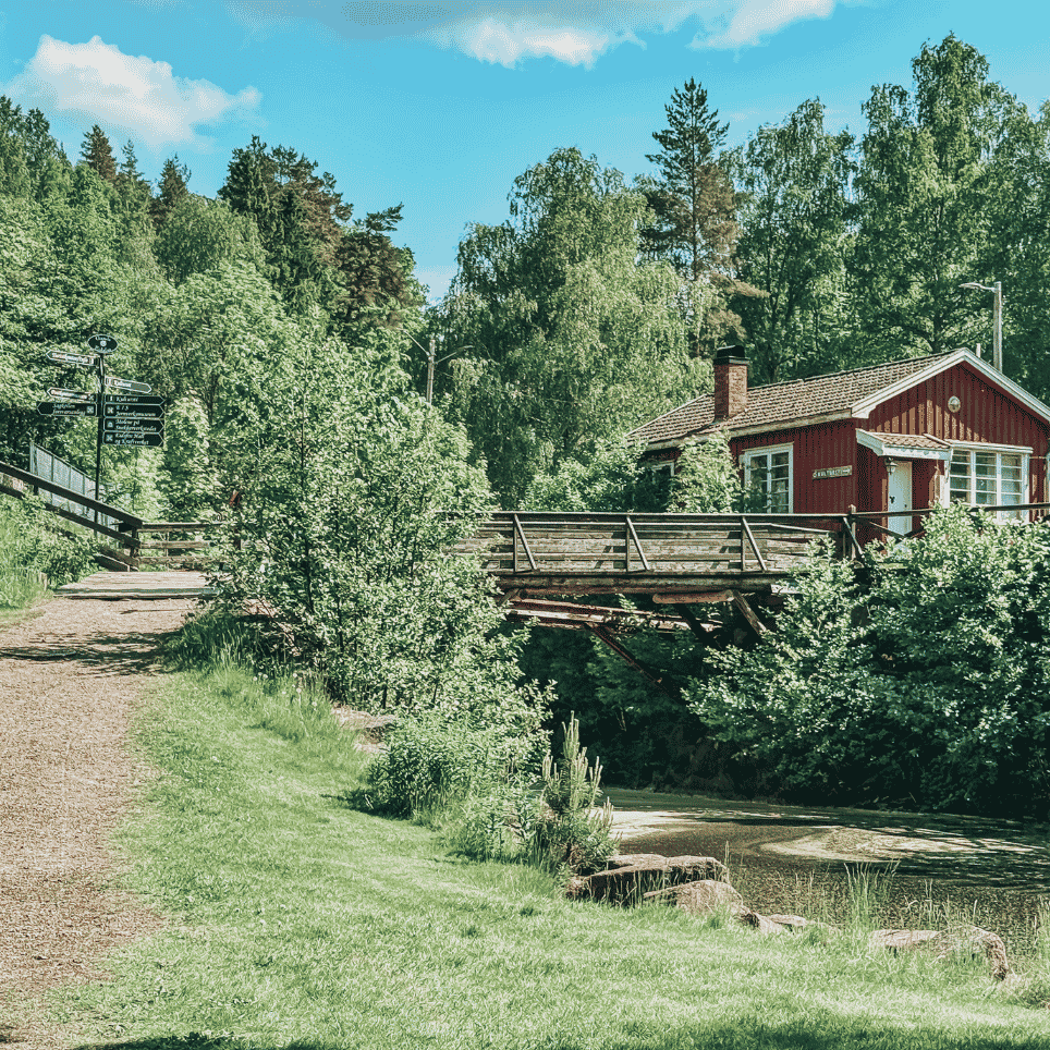 A small red cottage on a bridge over a stream, with a gravel path and a signpost.