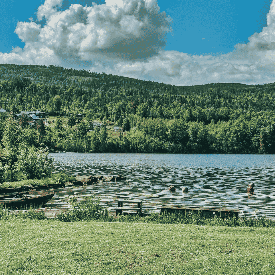 A view of a lake with a rowing boat, a lawn and wooded hills in the background.