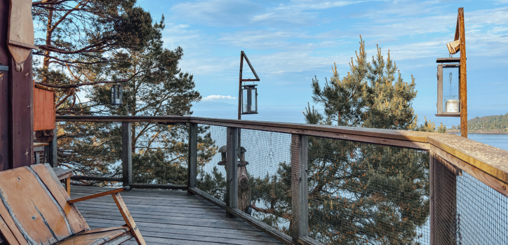 A wooden walkway running alongside a holiday cottage, with lanterns attached to the railing and a view of the sea.