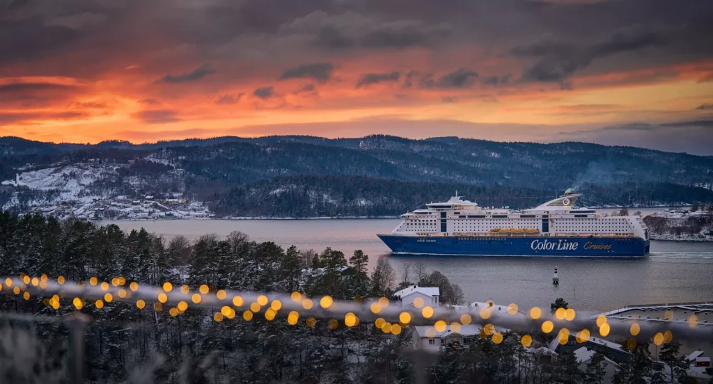 Color Fantasy fra Color Line seiler gjennom en snødekt fjord i solnedgangen, med en fargerik himmel og opplyste julelys i forgrunnen.